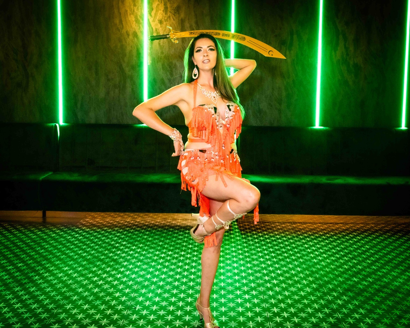 Lizzie balancing a sword on her head in a coral fringed costume, lit by green neon